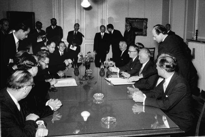 Adenauer unterzeichnet das Luxemburger Abkommen an einem großen, runden Tisch, an dem viele Männer sitzen