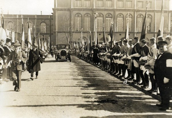 Schwarz-Weiss-Aufnahme von einem Automobil auf einer Straße, umgeben von Soldaten mit Flaggen