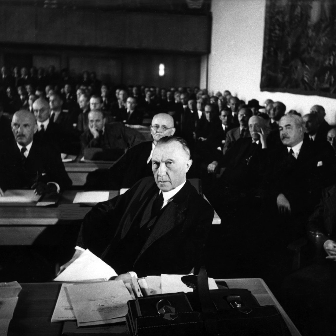 Konrad Adnenauer sitzt am Tisch, hinter ihm sitzen noch mehr Personen