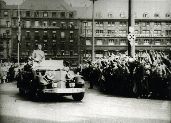 Schwarz-Weiss-Aufnahme von Adolf Hitler in einem Automobil während ihm die umstehenden Menschen den Hitlergruß zeigen 
