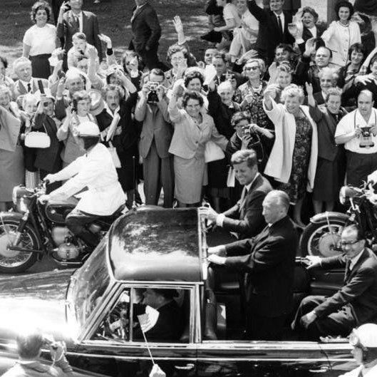 Adenauer und Kennedy sitzen in einem offenen Auto. Die Menschen am Straßenrand jubeln ihnen zu.