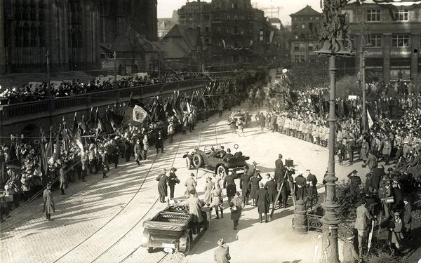 Schwarz-Weiss-Aufnahme von einem Straßenzug mit zahlreichen Menschen und zwei Automobilen