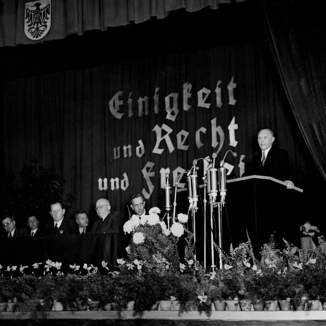 Adenauer vor einem Rednerpult und hinter ihm Schrift "Einigkeit und Recht und Freiheit"