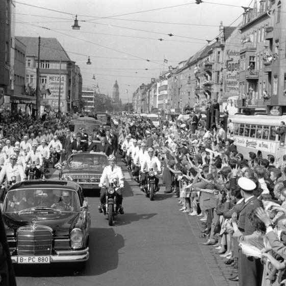 Eine Autoparade zieht an vielen jubelnden Menschen vorbei. Im mittleren, offenen Auto sitzen John F. Kennedy und Konrad Adenauer.