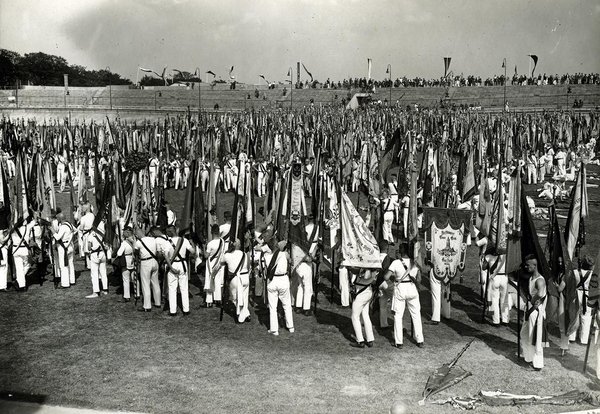 Schwarz-Weiss-Aufnahme von zahlreichen Männern in weißer Kleidung und mit Flaggen auf einem Sportplatz