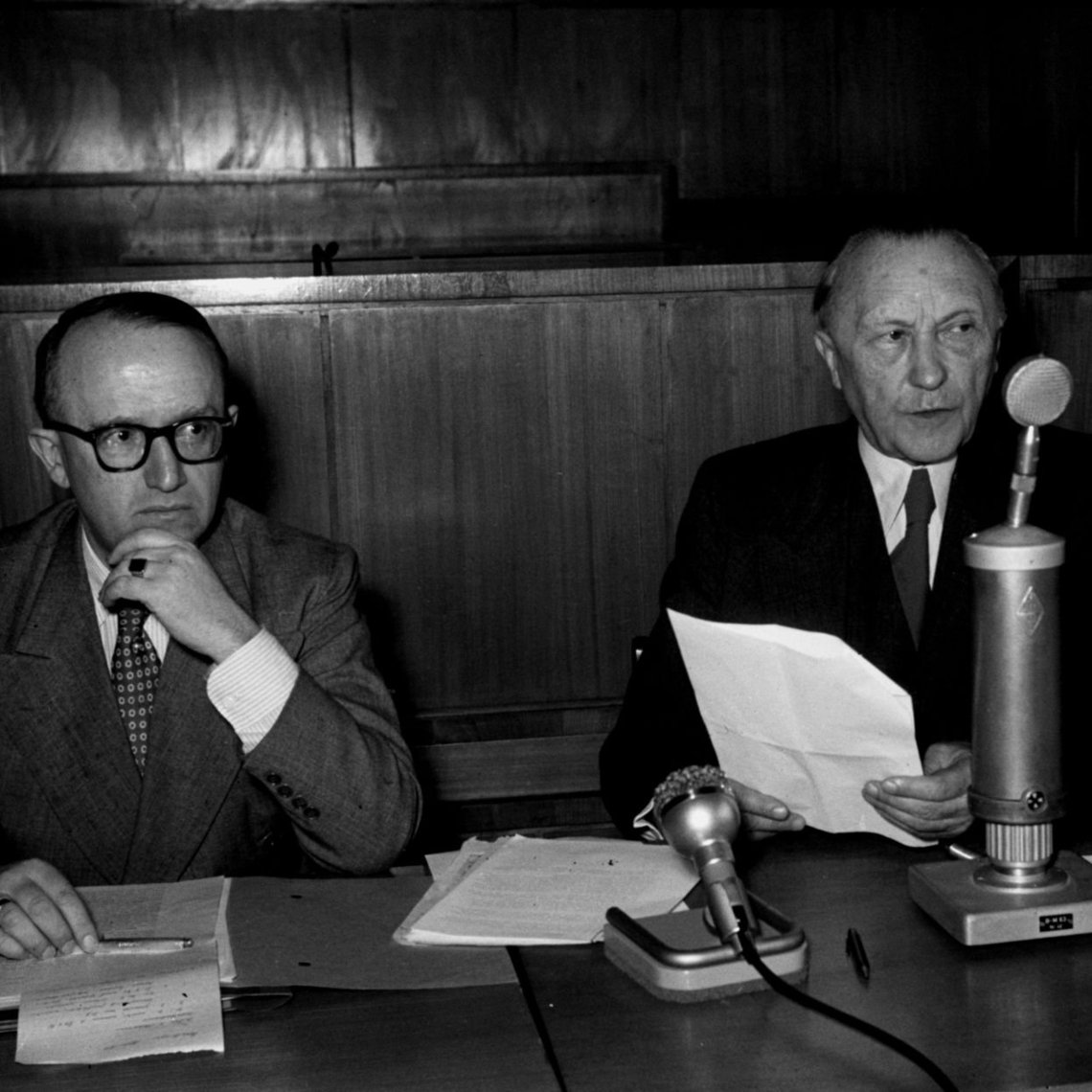 Konrad Adenauer sitzt mit Unterlagen am Mikrofon neben Walter Hallstein während einer Pressekonferenz