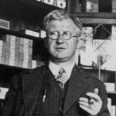 Wilhelm Elfes vor einem Regal mit Brille und Zigarre in der Hand, redend