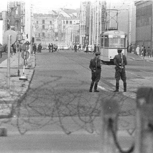 Im Vordergrund ist ein Stacheldrahtzaun zu sehen, dahinter zwei Polizisten die Wache stehen. Im Hintergrund ist Berlin zu sehen.