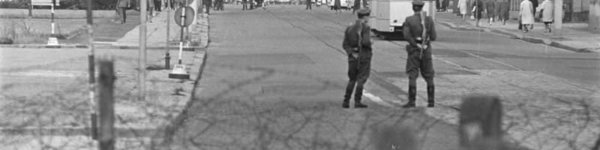 Im Vordergrund ist ein Stacheldrahtzaun zu sehen, dahinter zwei Polizisten die Wache stehen. Im Hintergrund ist Berlin zu sehen.