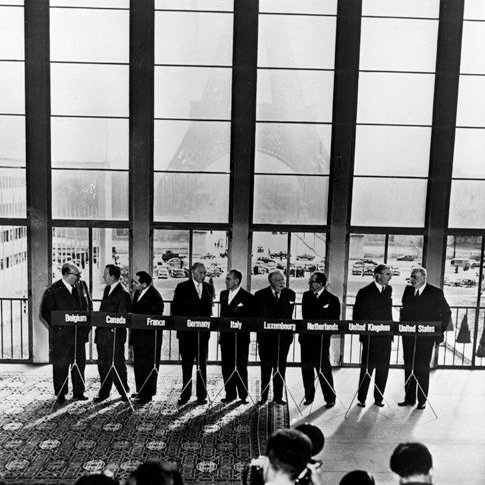 Konrad Adenauer steht mit den Vertretern der Gründermitglieder der Westeuropäischen Union während der Pariser Konferenzen an Rednerpulten. Im Hintergrund ist der Eifelturm zu sehen.
