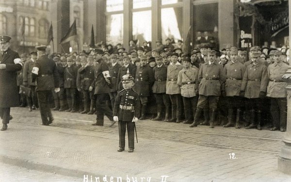Schwarz-Weiss-Aufnahme von einem kleinen Jungen in Soldatenuniform vor Männern in Reihenaufstellung
